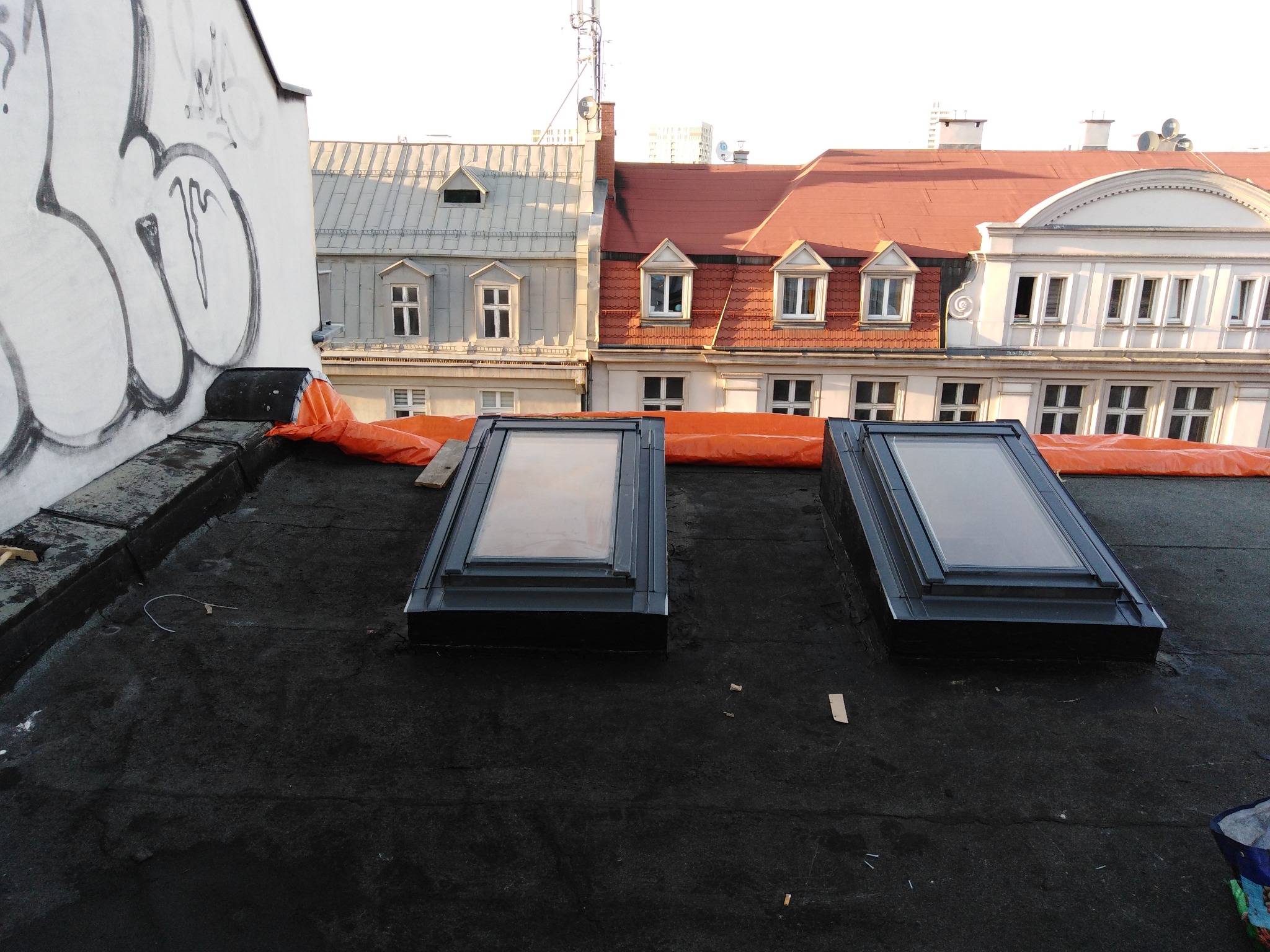 Dwa okna dachowe zamontowane na czarnym, płaskim dachu pokrytym papą, z widoczną pomarańczową folią uszczelniającą i budynkami w tle.