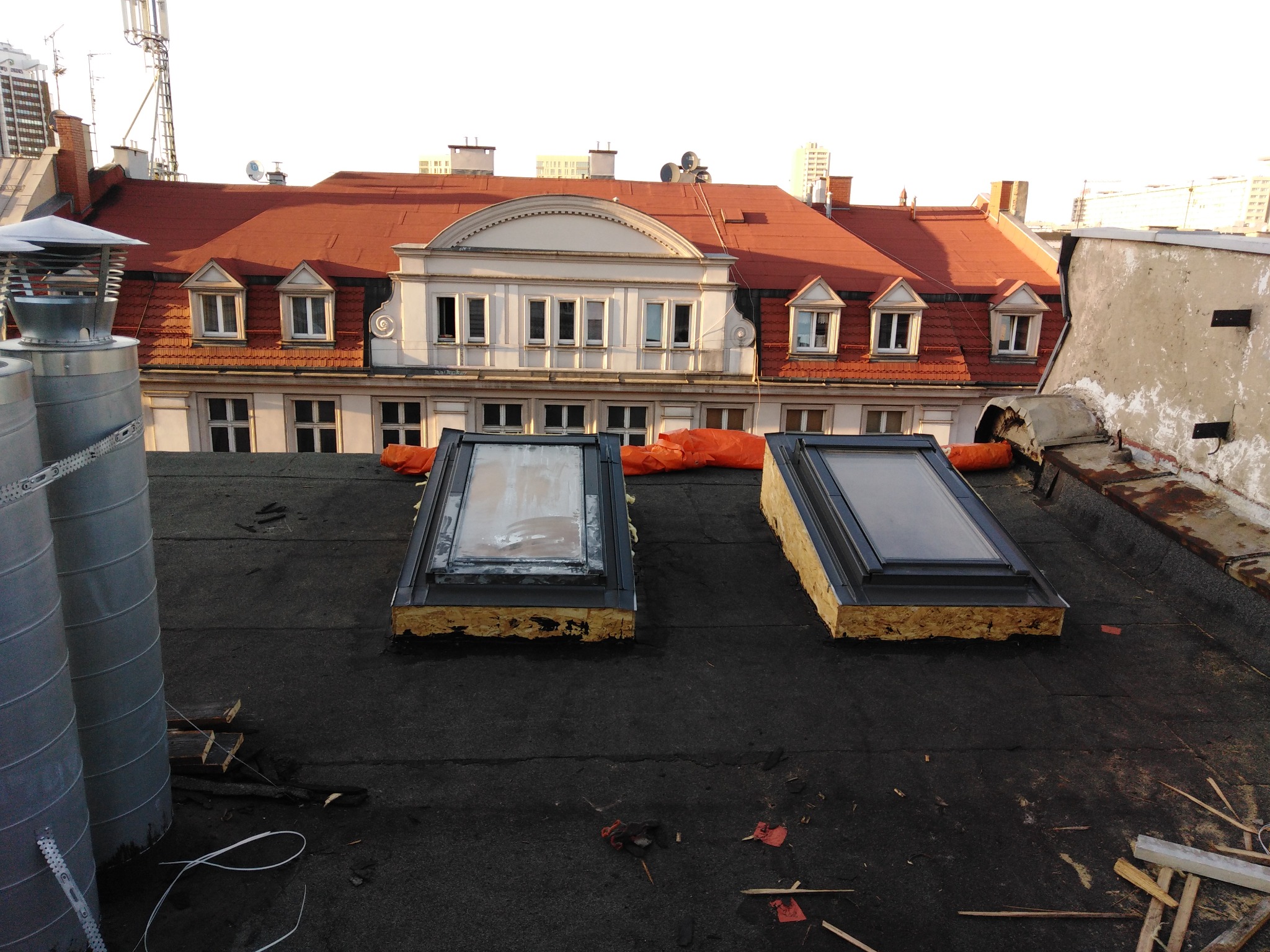 Dwa okna dachowe z czarnymi ramami, zamontowane na czarnym pokryciu dachowym z papy, w tle budynki miejskie i komin wentylacyjny.