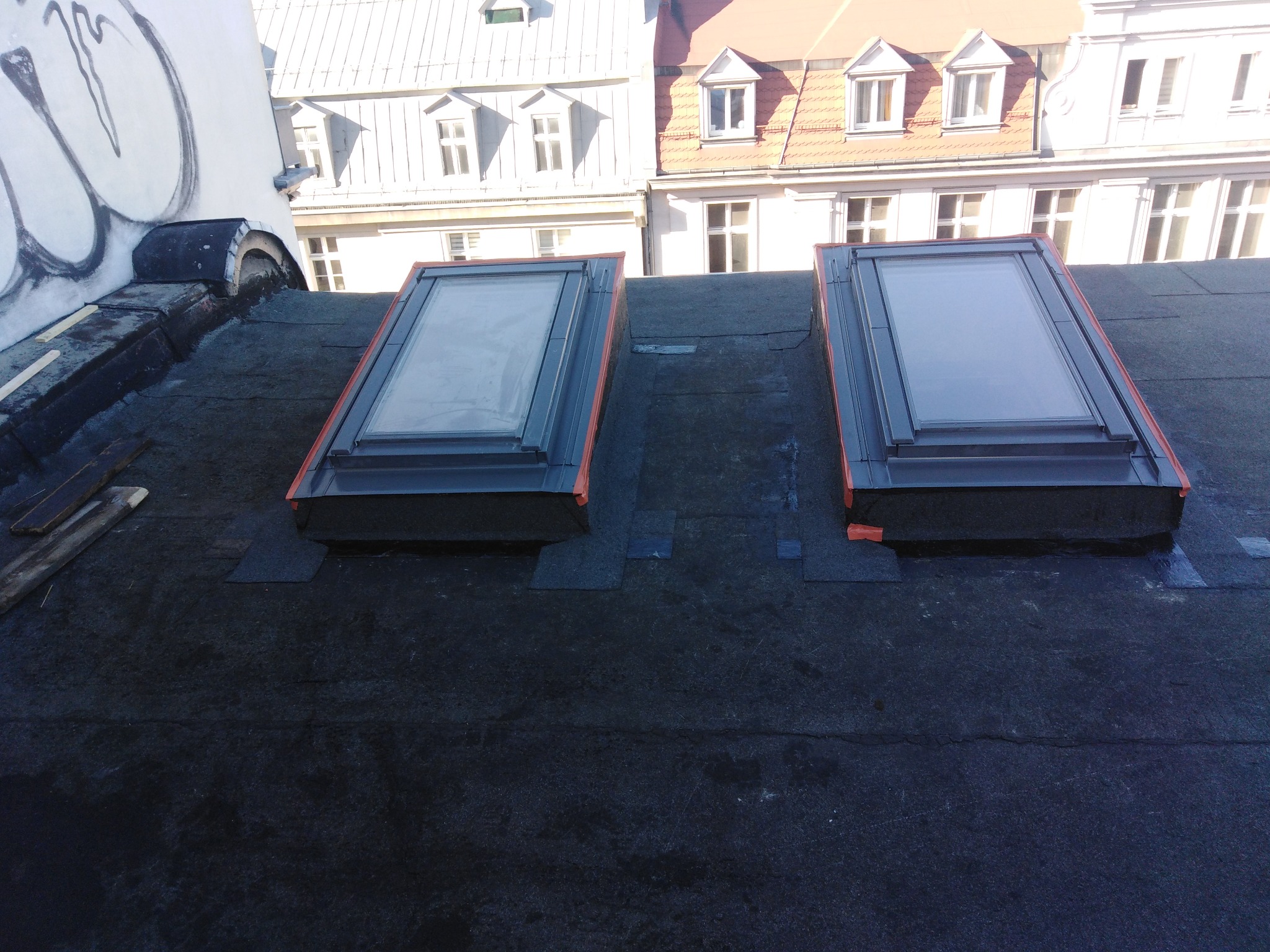 Dwa okna dachowe zamontowane na płaskim dachu pokrytym papą, widoczne budynki w tle.