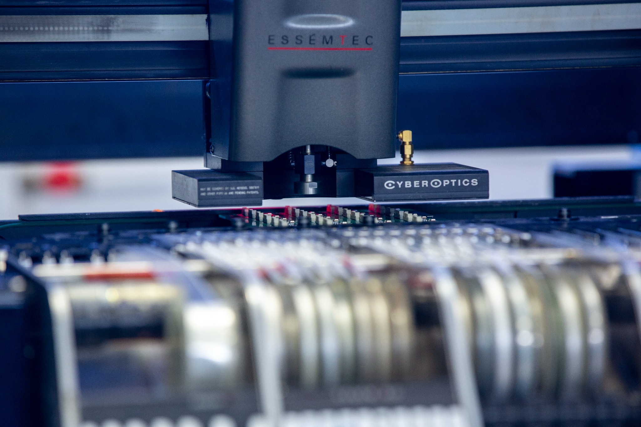 Precyzyjny automat przemysłowy marki Essemtec z modułem CyberOptics umieszczający komponenty elektroniczne na płytce drukowanej, widoczne podajniki komponentów.