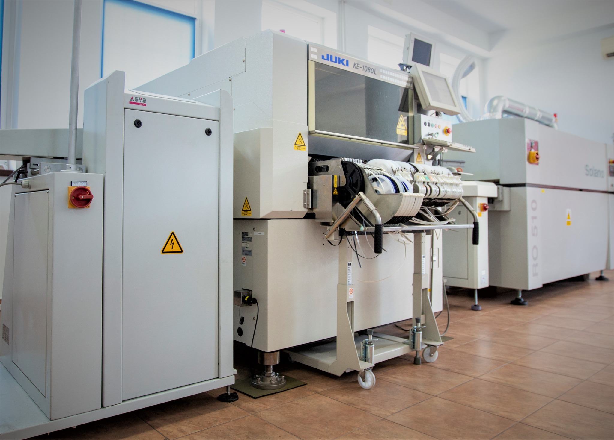 Profesjonalna linia produkcyjna JUKI KE-1080L i Solano RO 510 w jasnym pomieszczeniu, widoczne znaki ostrzegawcze.