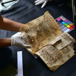 Otwarta, zniszczona księga z odręcznym pismem, trzymana w białych rękawiczkach na czarnym tle, obok karta kalibracyjna kolor&oacute;w.