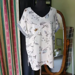 Bluzka z krótkim rękawem w kwiatowy wzór na manekinie, widoczne zasłony w tle oraz owalne lustro, w odbiciu fragment stołu krawieckiego z maszynami do szycia.