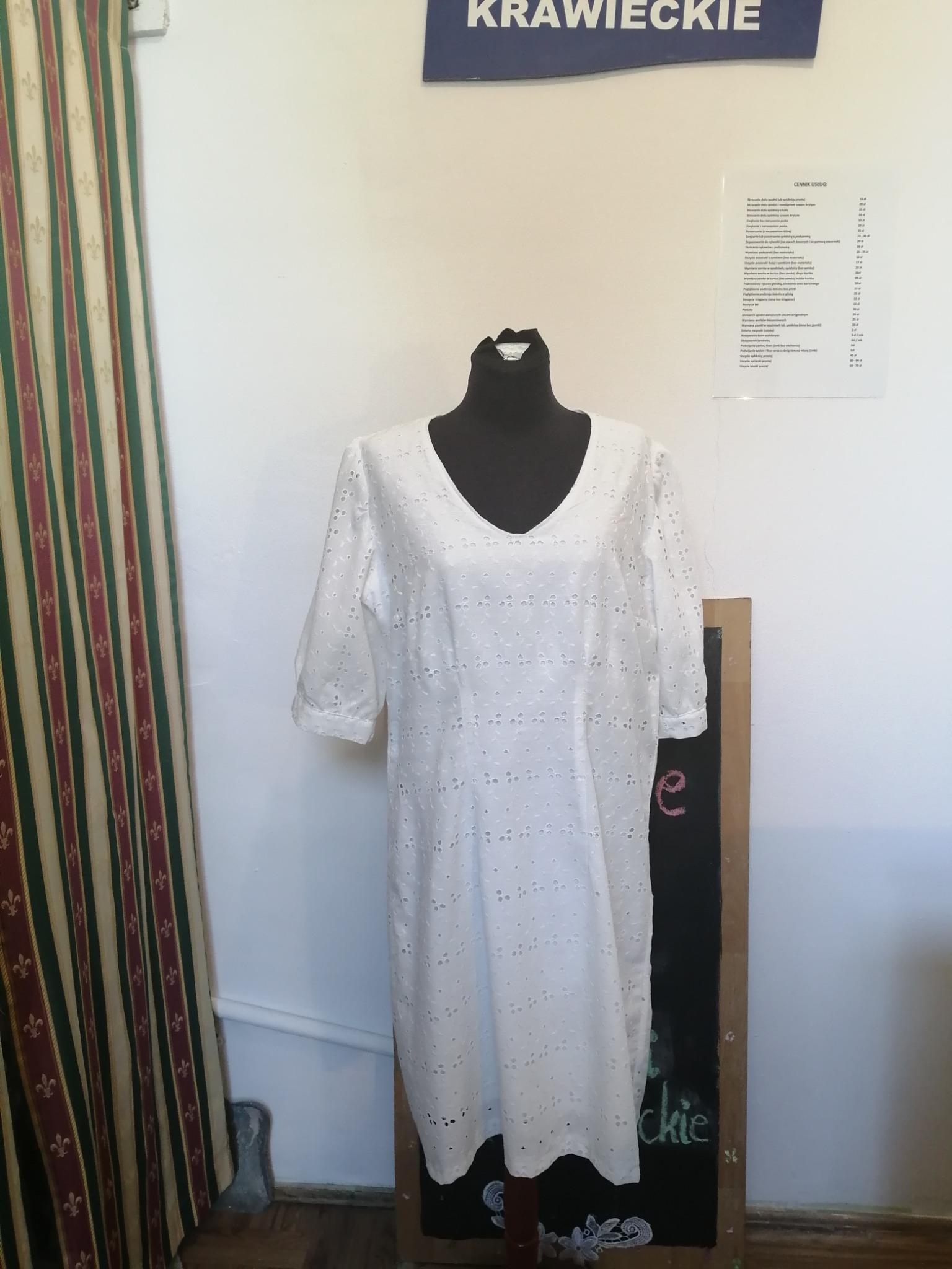 Biała, ażurowa sukienka na czarnym manekinie, widoczny fragment cennika usług krawieckich i zasłony w tle.