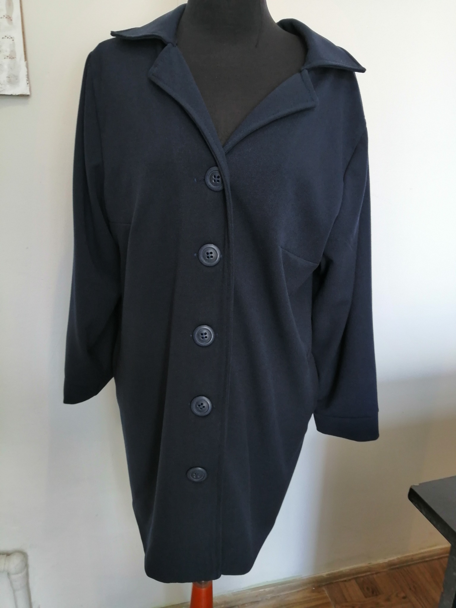 Granatowy płaszcz damski z kołnierzem i rzędem dużych guzików, prezentowany na manekinie w pracowni krawieckiej.