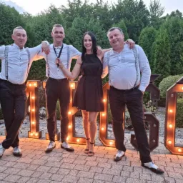 Czteroosobowa grupa w Oświęcimiu pozuje przed podświetlanymi literami tworzącymi napis, prawdopodobnie nazwę zespołu; mężczyźni w białych koszulach z szelkami i czarnych spodniach, kobieta...