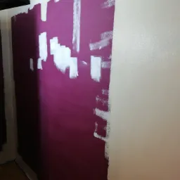 Ściana w trakcie przemalowywania z intensywnego fioletu na jasny beż, widoczne fragmenty białej farby gruntującej i taśmy malarskiej przy podłodze.