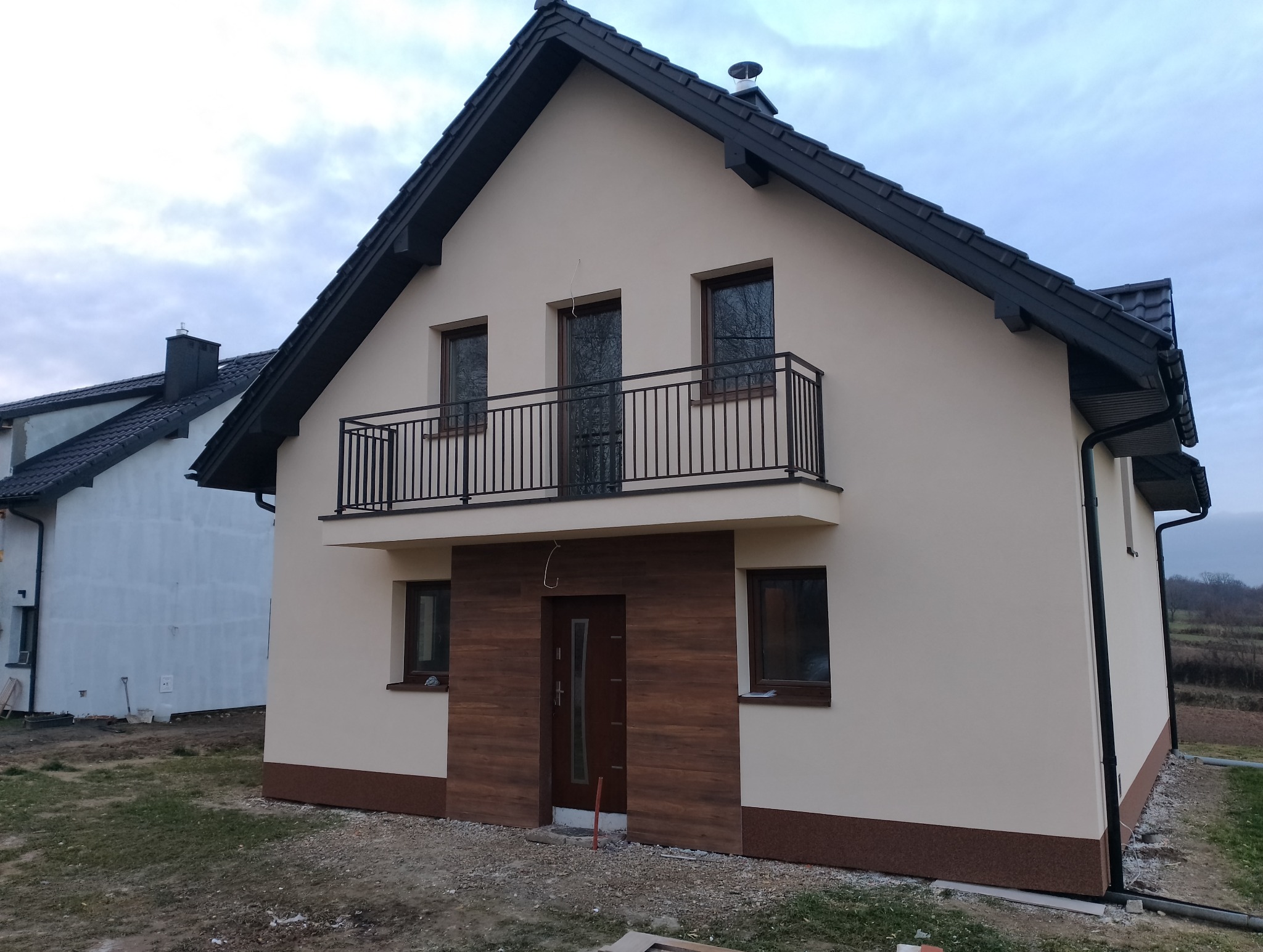 Nowoczesny dom jednorodzinny z beżową elewacją, czarnym dachem i balkonem z metalową balustradą, wykończony panelami imitującymi drewno wokół drzwi wejściowych.