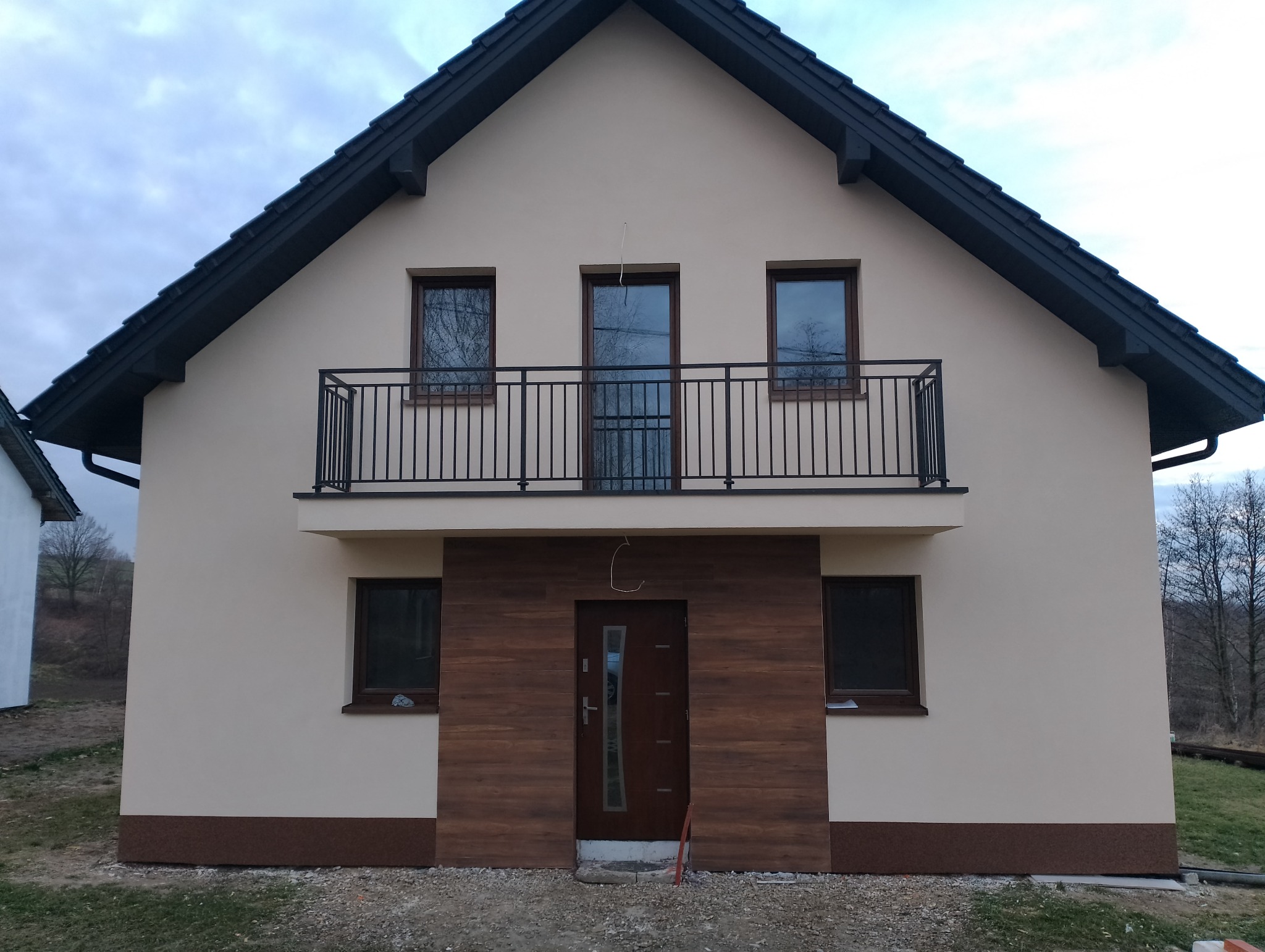 Nowoczesny dom z beżową elewacją, ciemnobrązową stolarką okienną i drewnopodobną okładziną wokół wejścia, balkon z czarną balustradą.