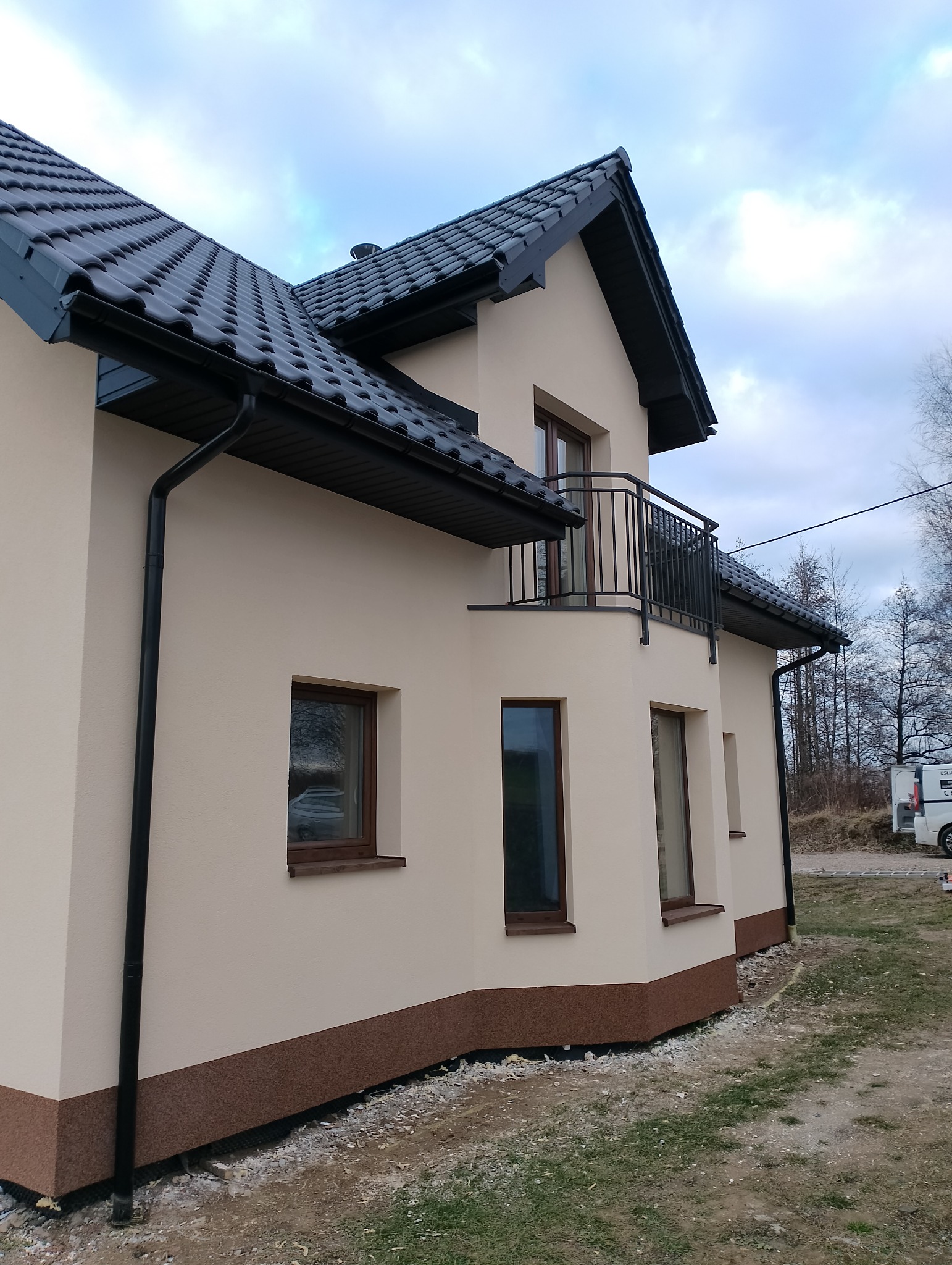 Dwukondygnacyjny dom z beżową elewacją, ciemnobrązowym cokołem i czarną, błyszczącą dachówką, widoczne rynny i balkon z metalową balustradą.