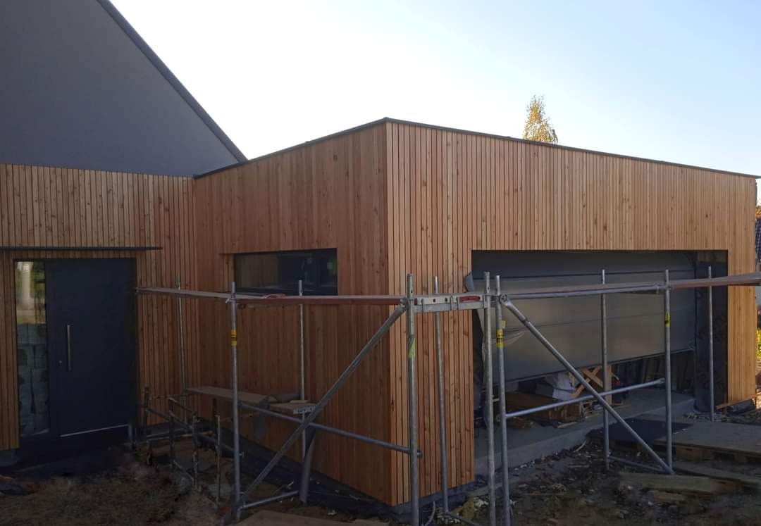 Nowoczesny dom z elewacją z jasnego drewna, widoczne rusztowania i częściowo otwarta brama garażowa, minimalistyczny design.