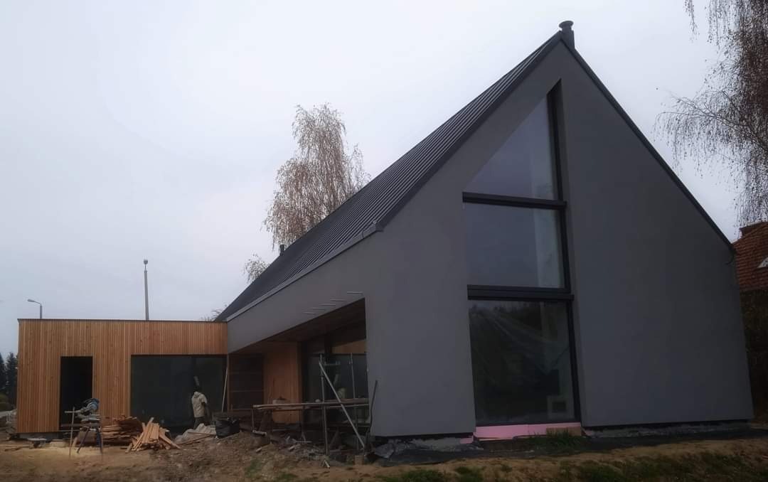 Nowoczesny dom jednorodzinny z szarą elewacją i drewnianą dobudówką w trakcie wykończenia, widoczne rusztowania i materiały budowlane.