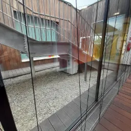 Widok przez szybę na zewnętrzną jednostkę klimatyzacji Daikin, umieszczoną pod metalowymi schodami z drewnianą balustradą, na tle ściany z drewnianych paneli i żwirowego podłoża.