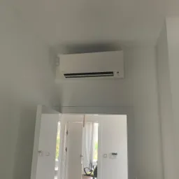 Biała jednostka klimatyzacji Daikin zamontowana na ścianie nad drzwiami w nowoczesnym, jasnym wnętrzu. Widoczny fragment biurka i krzesła w pokoju za drzwiami.