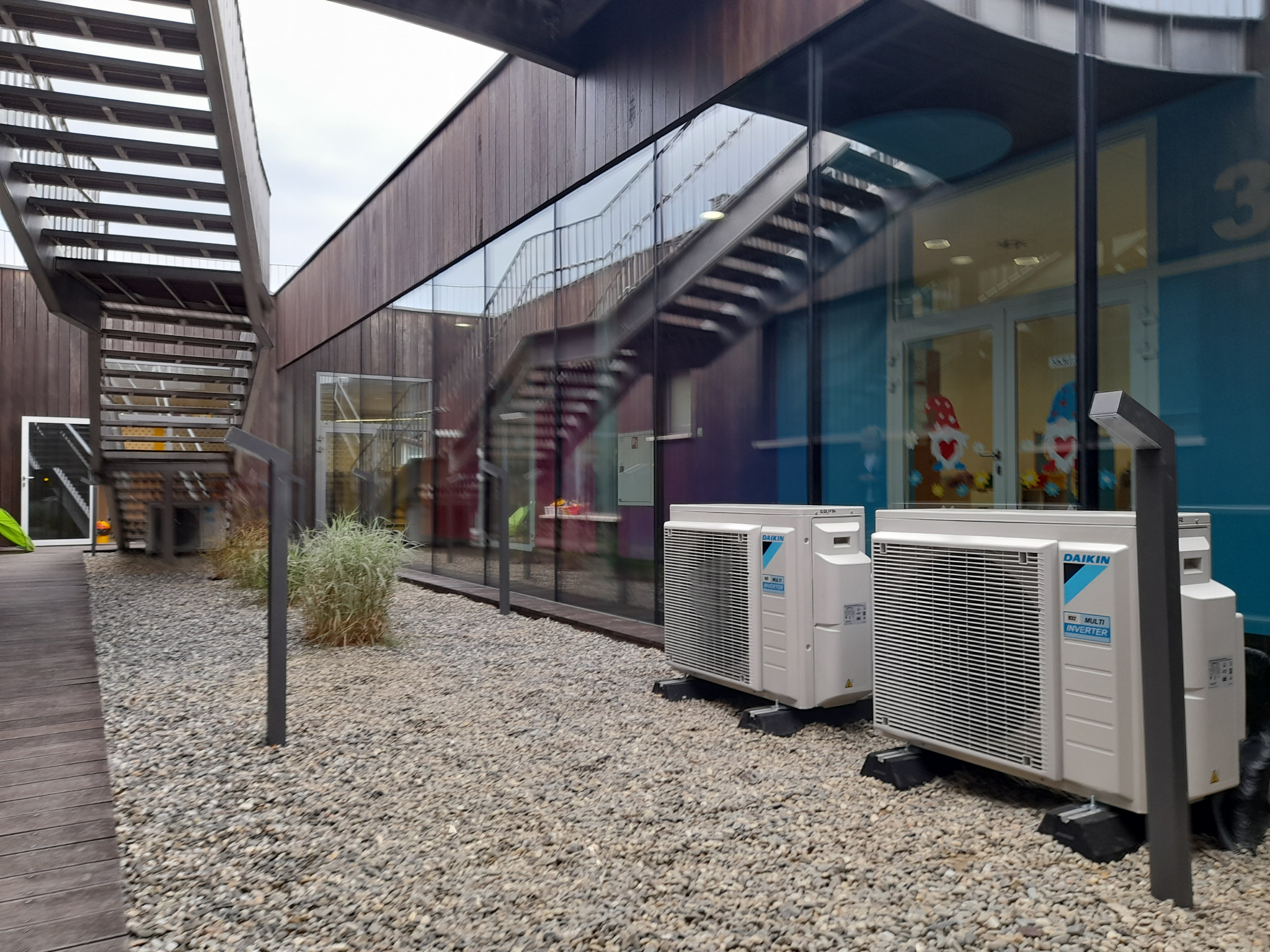 Dwa klimatyzatory Daikin Multi Inverter ustawione na żwirowym podłożu przed nowoczesnym budynkiem z drewnianą elewacją i szklanymi ścianami, z widocznymi schodami zewnętrznymi.