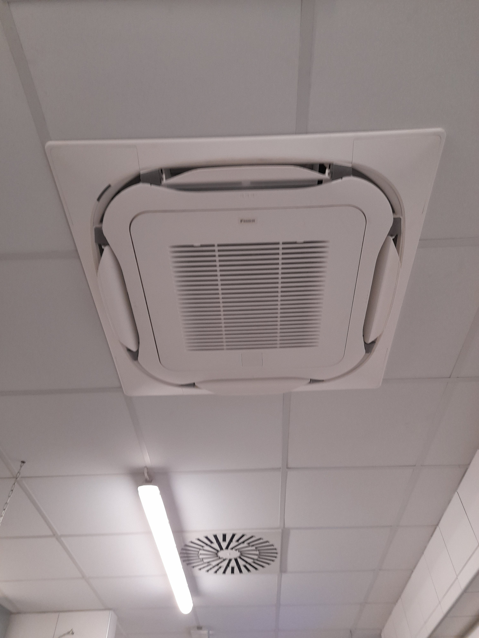 Kasetonowa jednostka klimatyzacyjna Daikin zamontowana w suficie podwieszanym biura, widoczny fragment oświetlenia i kratka wentylacyjna.