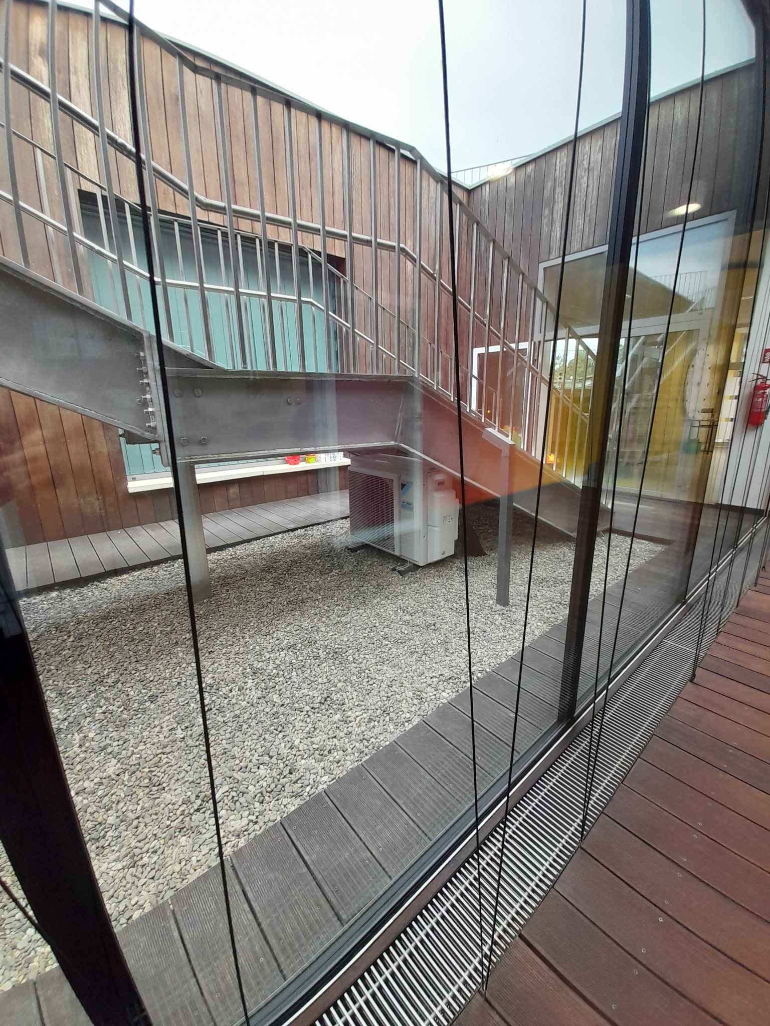 Widok przez szybę na zewnętrzną jednostkę klimatyzacji Daikin, umieszczoną pod metalowymi schodami z drewnianą balustradą, na tle ściany z drewnianych paneli i żwirowego podłoża.