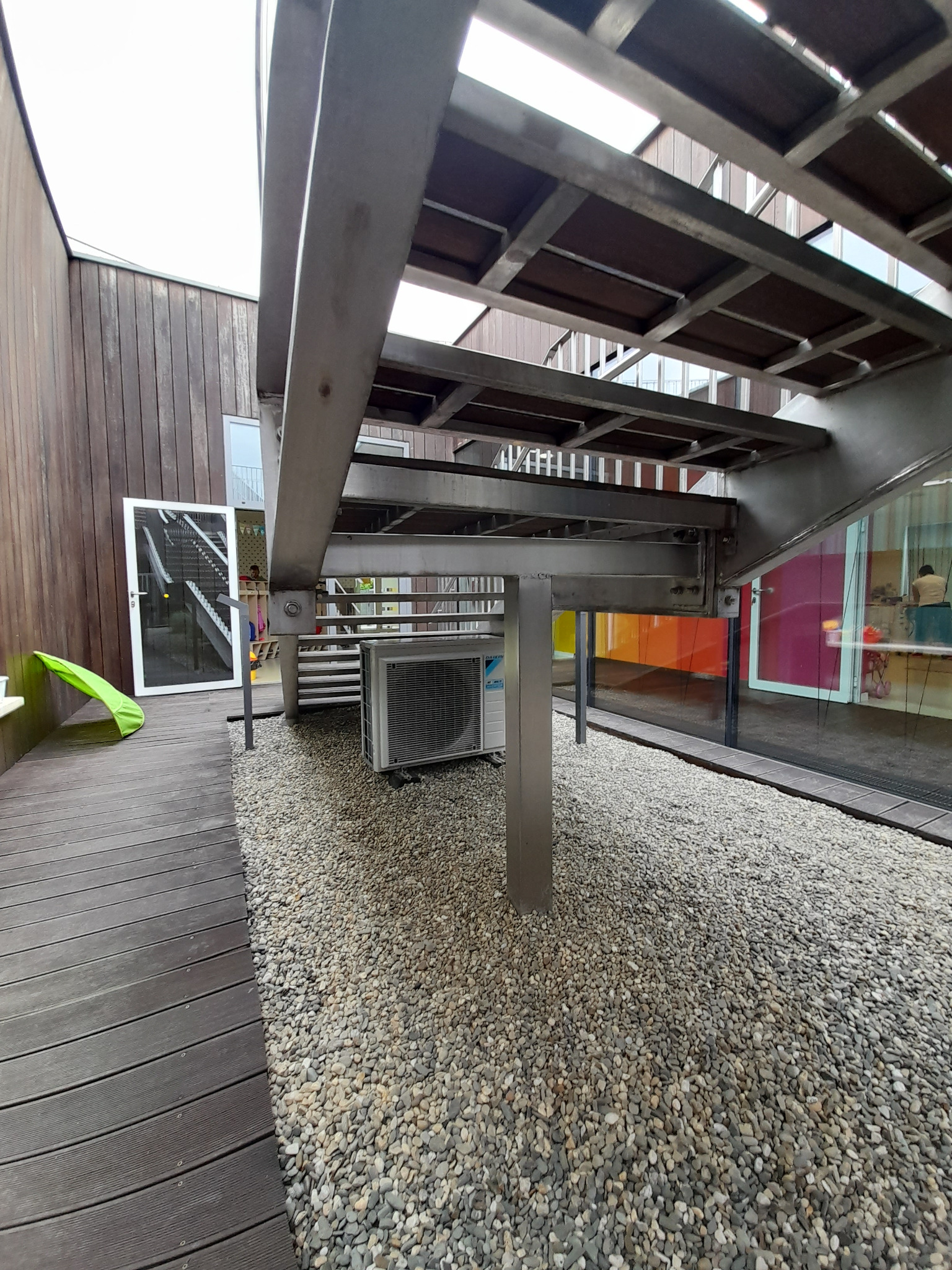 Zewnętrzna jednostka klimatyzacji Daikin pod metalowymi schodami, obok drewnianego tarasu i żwirowego podłoża, widok na kolorowe okna budynku w tle.