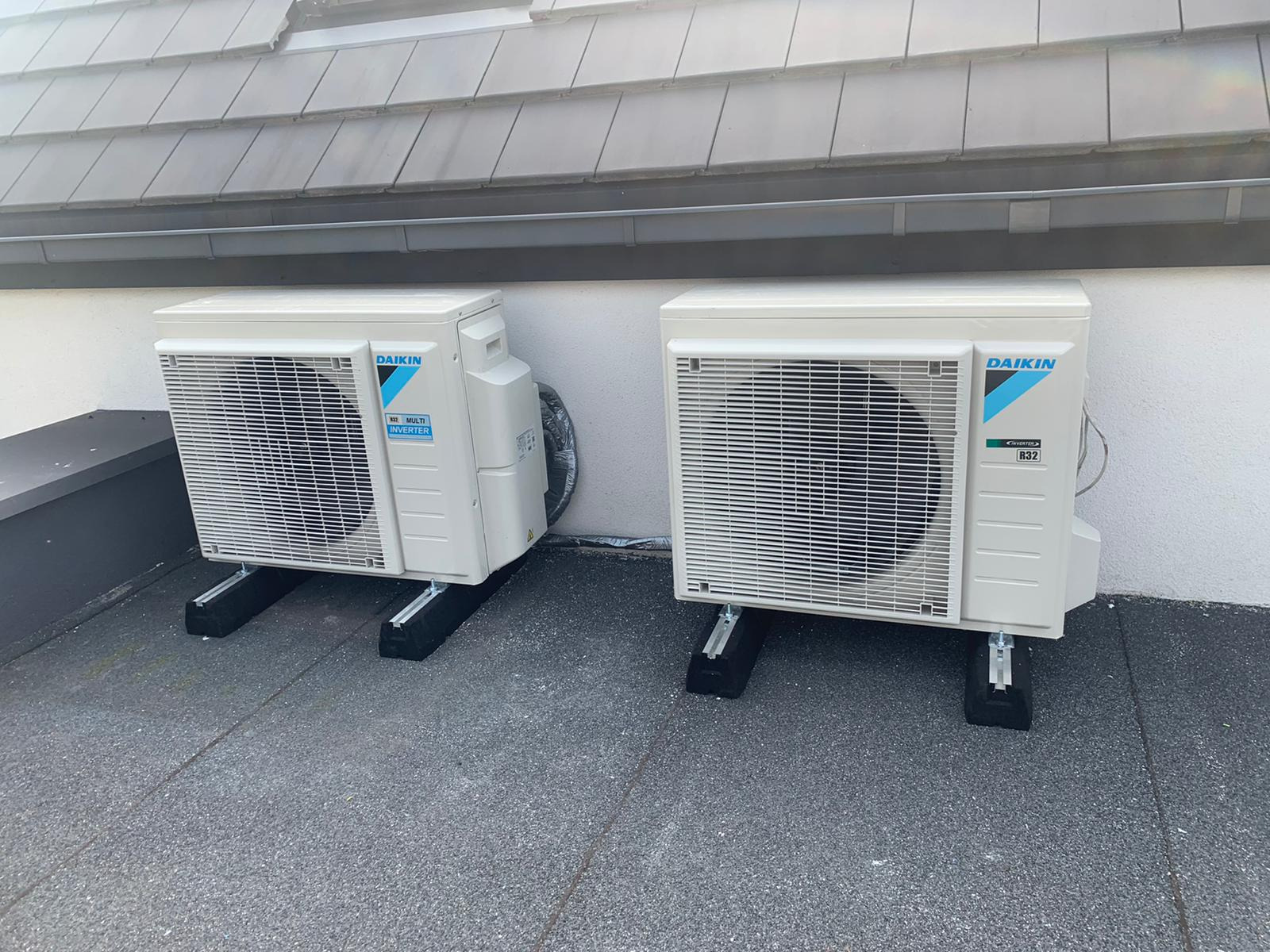 Dwa białe, zewnętrzne jednostki klimatyzacji Daikin Multi Inverter R32, zamontowane na czarnych, gumowych podstawach na szarym, płaskim dachu z widoczną krawędzią dachu i szarą ścianą budynku w tle.