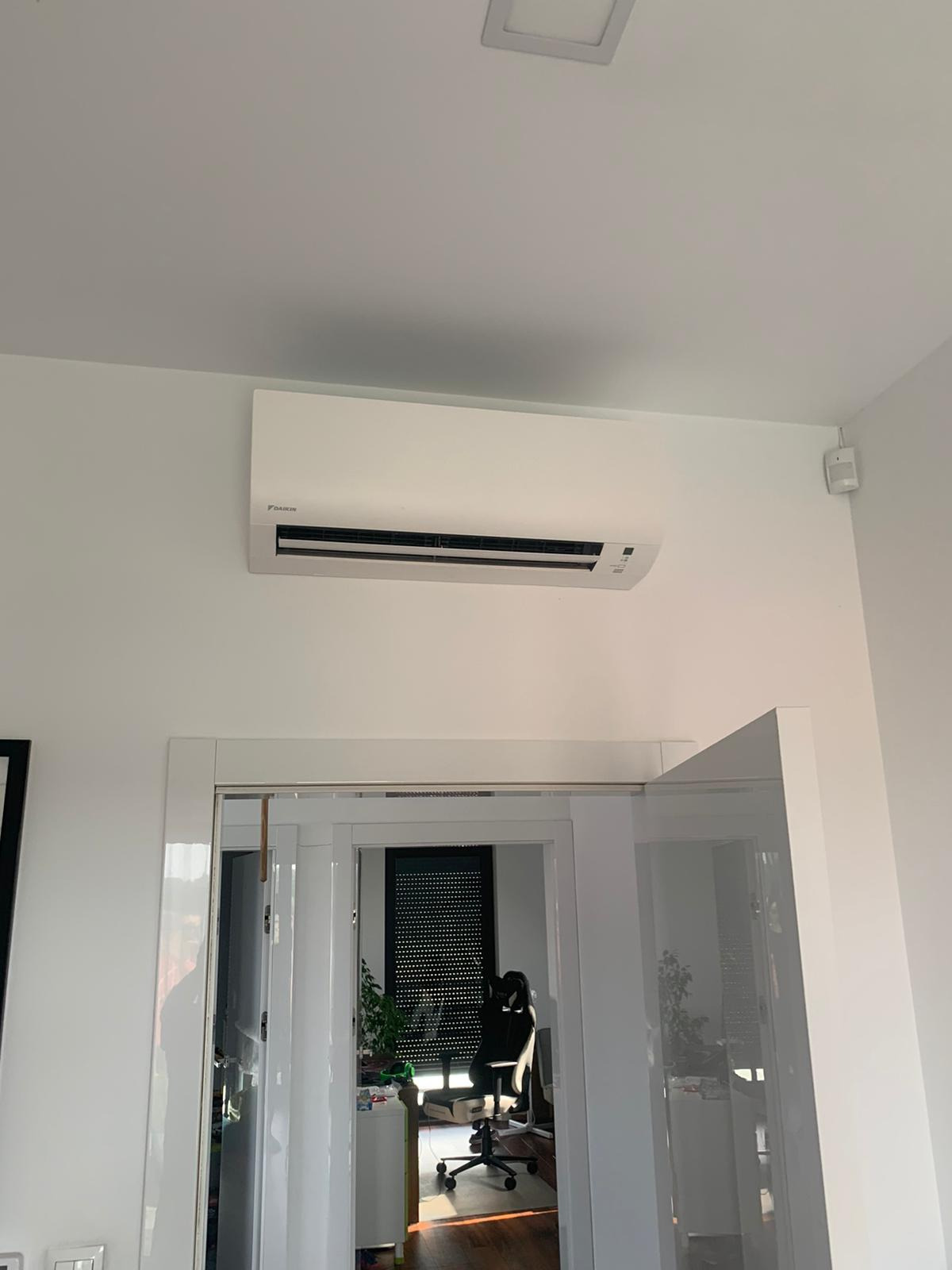 Biała jednostka klimatyzacyjna Daikin zamontowana na ścianie nad przeszklonymi drzwiami przesuwnymi w nowoczesnym wnętrzu. Widoczny czujnik ruchu na ścianie w prawym górnym rogu.