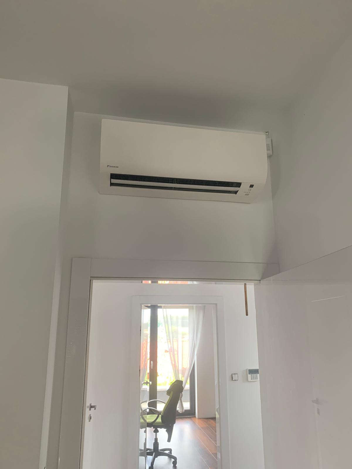 Biały klimatyzator Daikin zamontowany na ścianie nad białymi drzwiami, widok z korytarza na jasny pokój z biurkiem i krzesłem.
