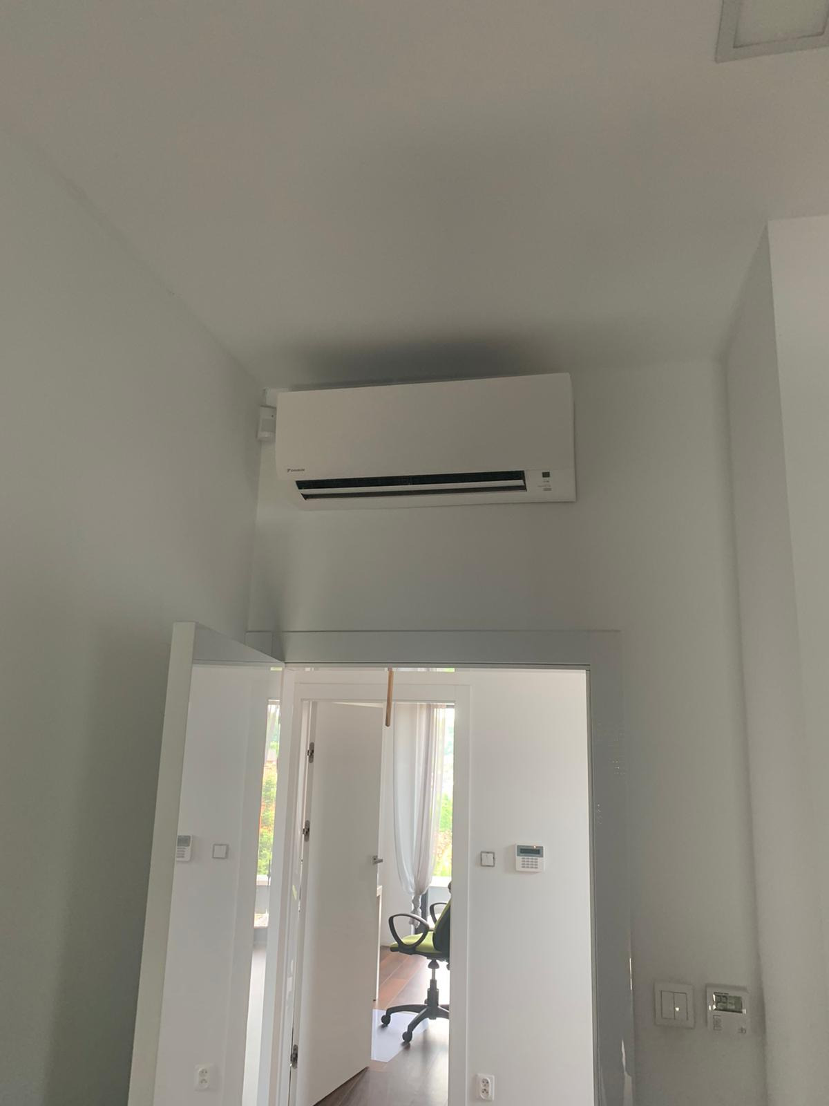 Biała jednostka klimatyzacji Daikin zamontowana na ścianie nad drzwiami w nowoczesnym, jasnym wnętrzu. Widoczny fragment biurka i krzesła w pokoju za drzwiami.