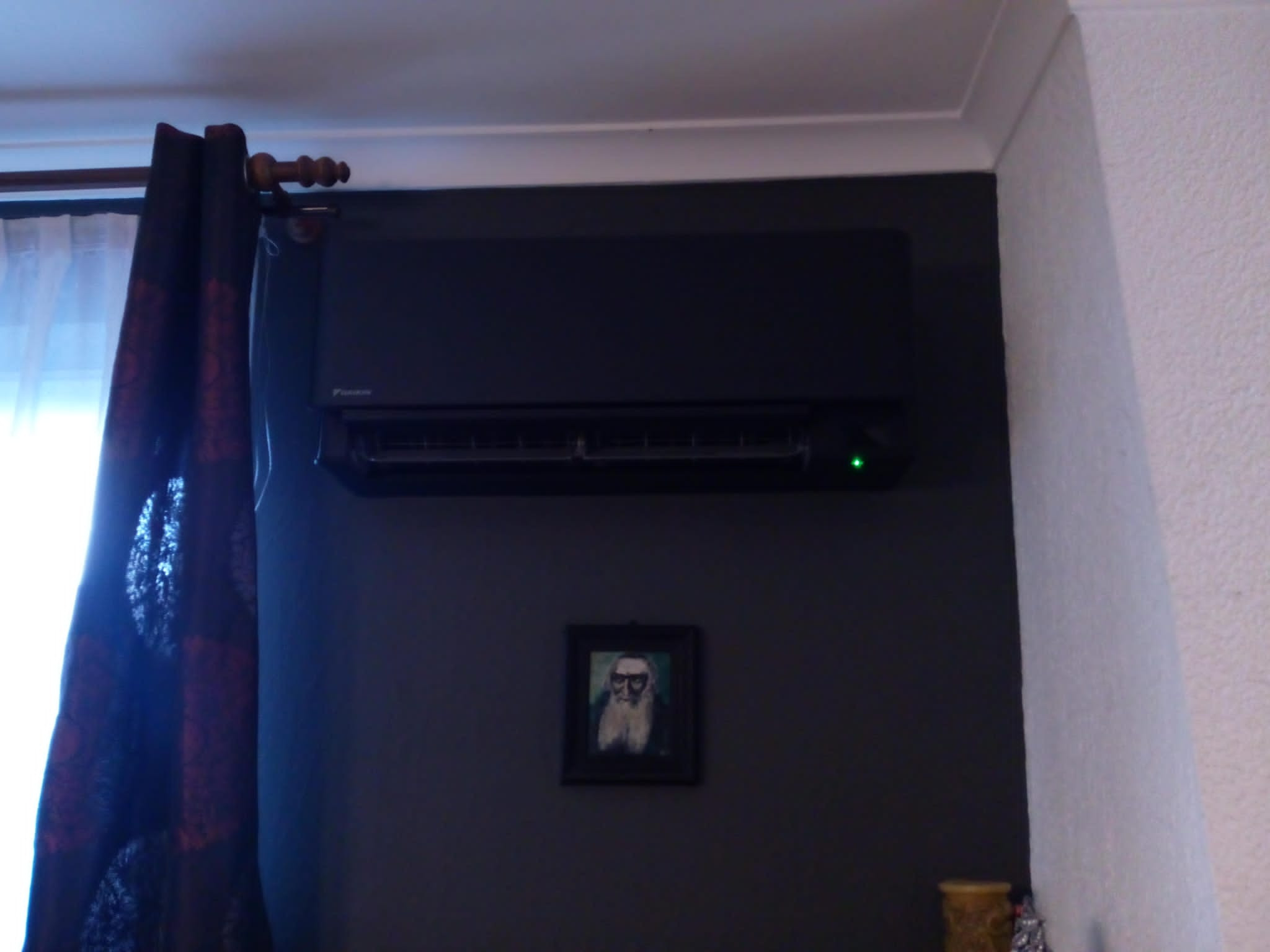 Czarna klimatyzacja Daikin zamontowana na ciemnoszarej ścianie, pod nią wisi obrazek w ramce, z boku zasłonięte okno.