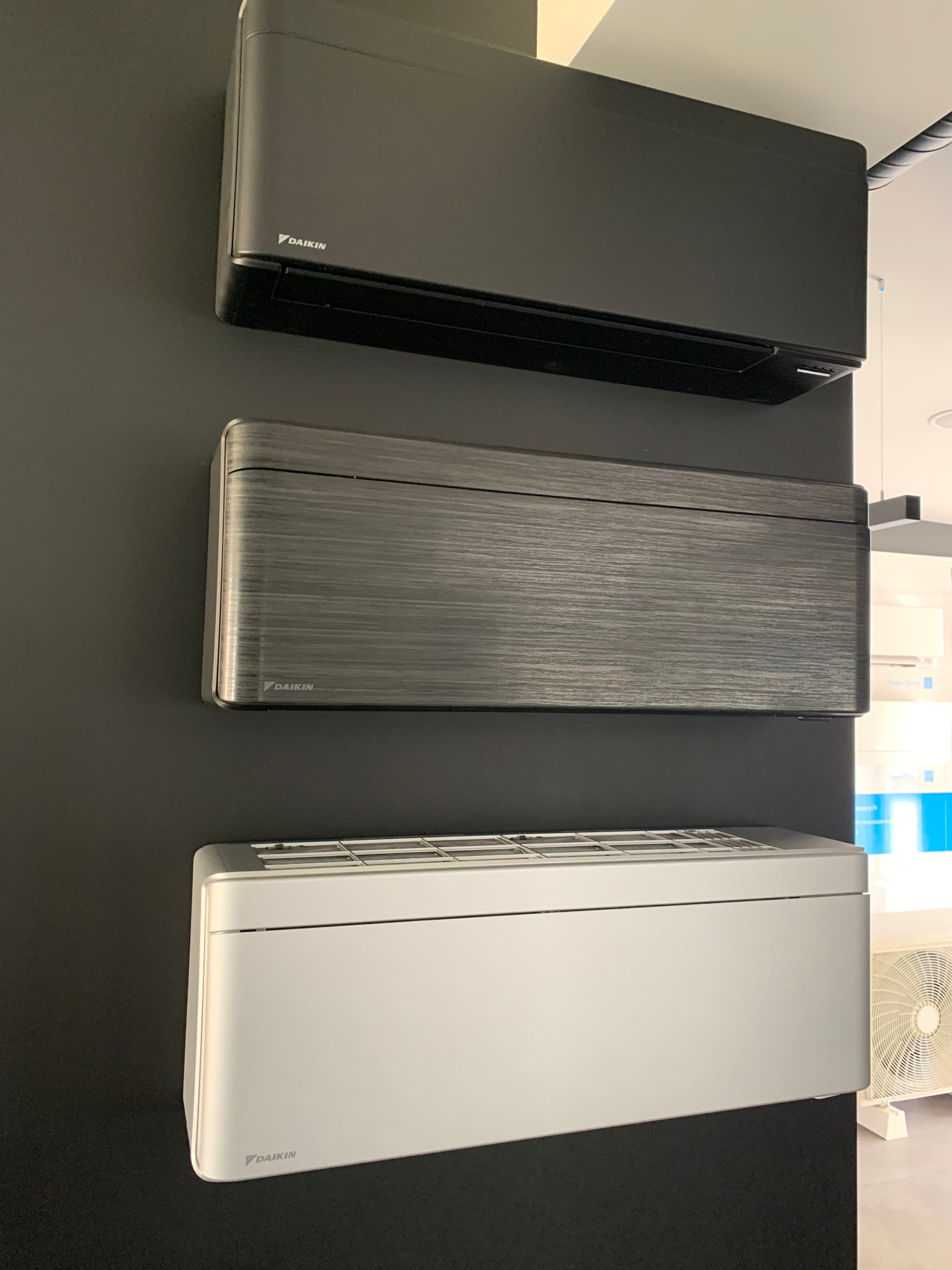 Trzy jednostki wewnętrzne klimatyzacji Daikin w różnych kolorach (czarny, grafitowy, srebrny) zamontowane na ciemnej ścianie. Widoczny fragment klimatyzatora stojącego.