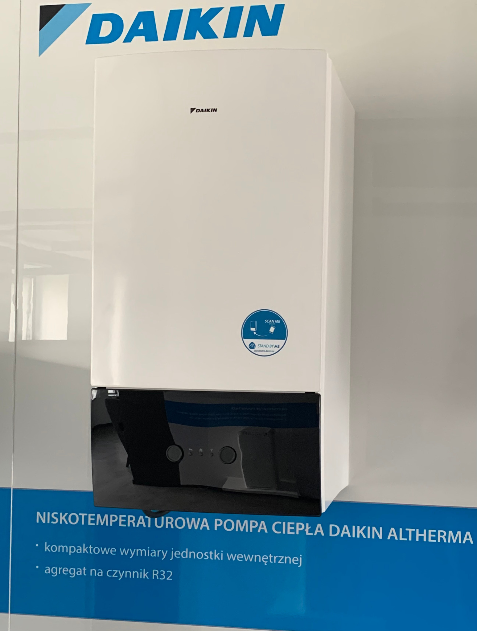 Biała jednostka wewnętrzna niskotemperaturowej pompy ciepła Daikin Altherma, widoczna na tle ściany z logo Daikin i informacjami o produkcie, prezentowana w pomieszczeniu.