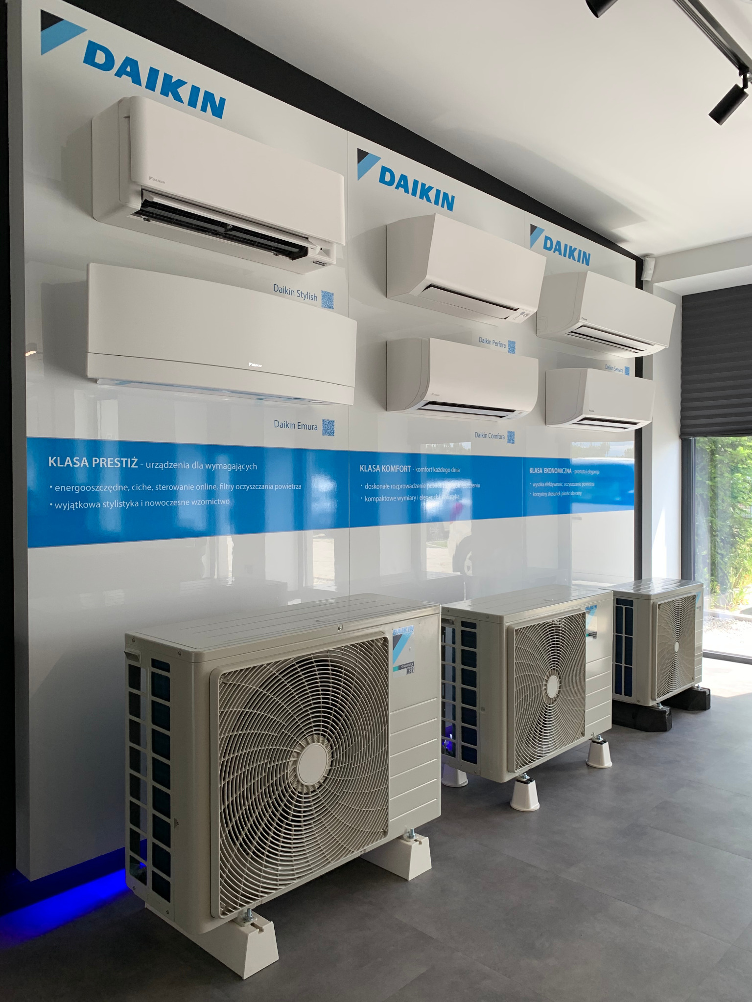 Ekspozycja klimatyzatorów Daikin w salonie, prezentująca modele ścienne i zewnętrzne jednostki, z widocznymi tablicami informacyjnymi o klasach i cechach urządzeń.