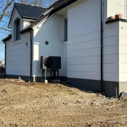 Czarna pompa ciepła LG na betonowych podstawach, zamontowana przy białej ścianie domu jednorodzinnego z czarną dachówką, widok z poziomu gruntu na nierówny, brązowy teren.