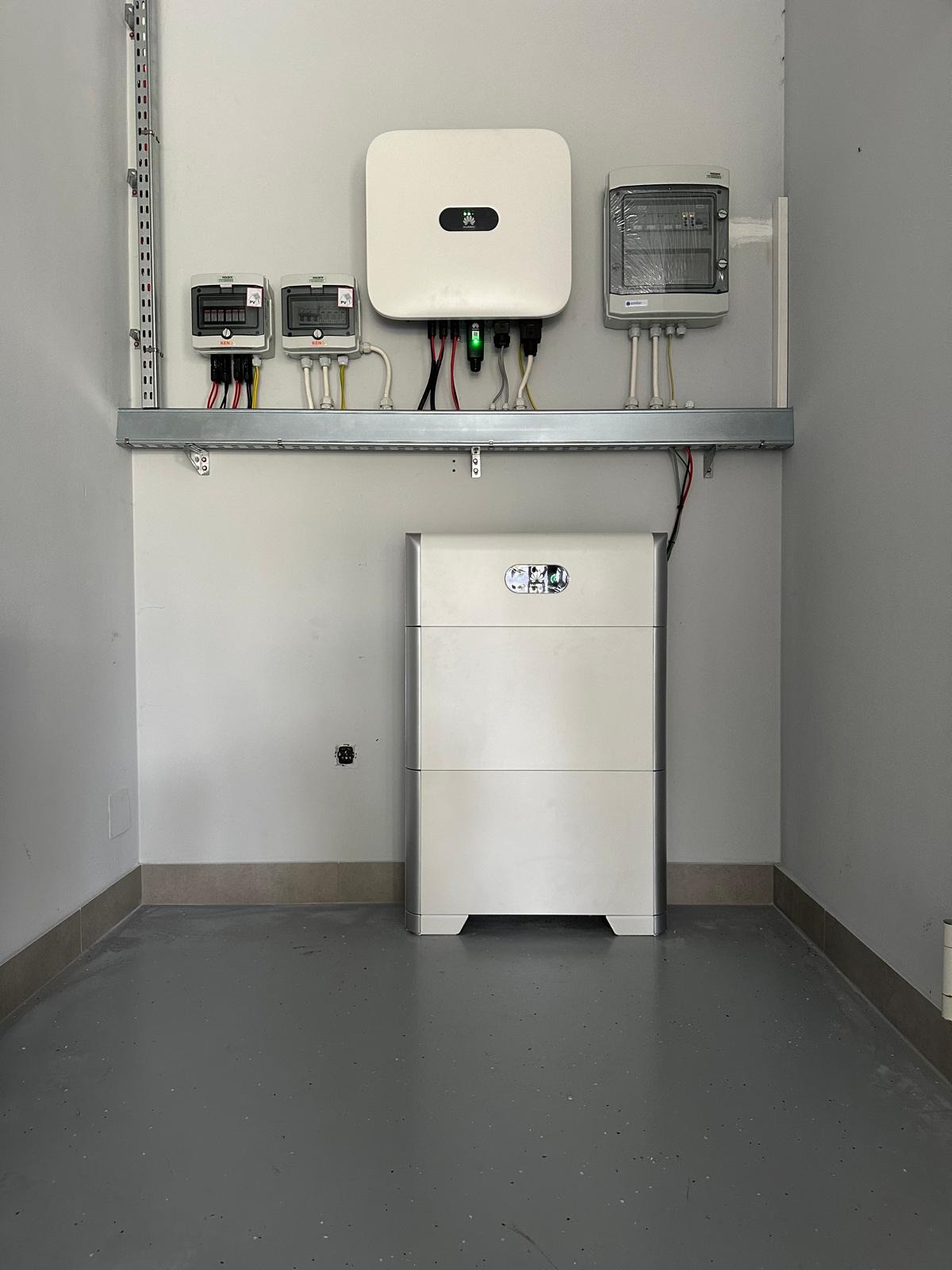 Biały akumulator energii z falownikiem i licznikami na ścianie w szarym pomieszczeniu technicznym. Widoczne przewody i elementy instalacji elektrycznej.