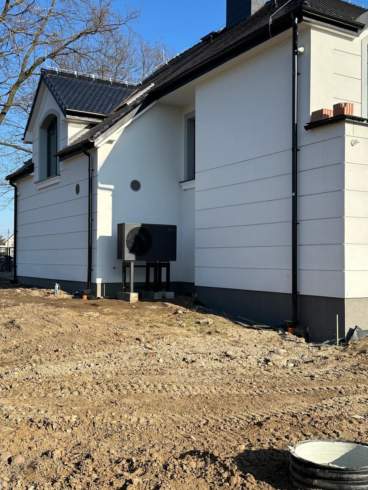 Czarna pompa ciepła LG na betonowych podstawach, zamontowana przy białej ścianie domu jednorodzinnego z czarną dachówką, widok z poziomu gruntu na nierówny, brązowy teren.