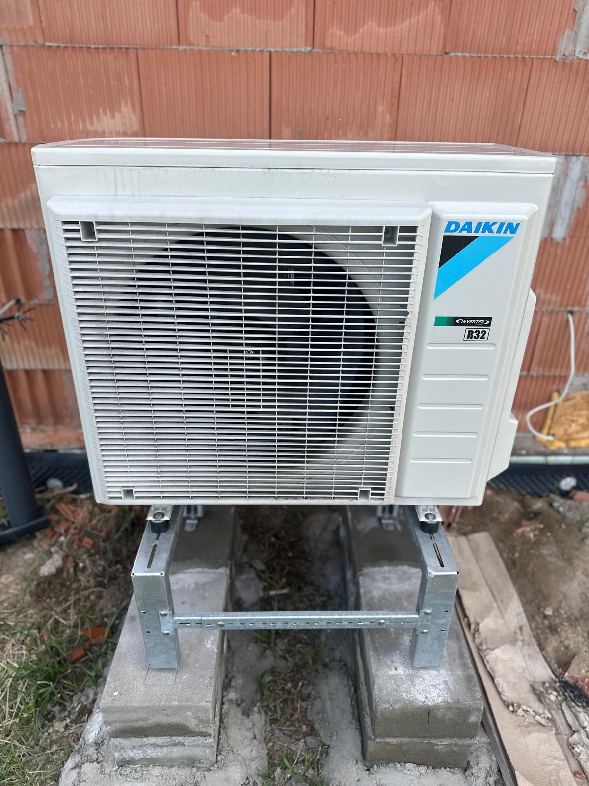 Zewnętrzna jednostka klimatyzacji Daikin na metalowej konstrukcji wsporczej, zamontowana na betonowych bloczkach przed murem z czerwonej cegły.