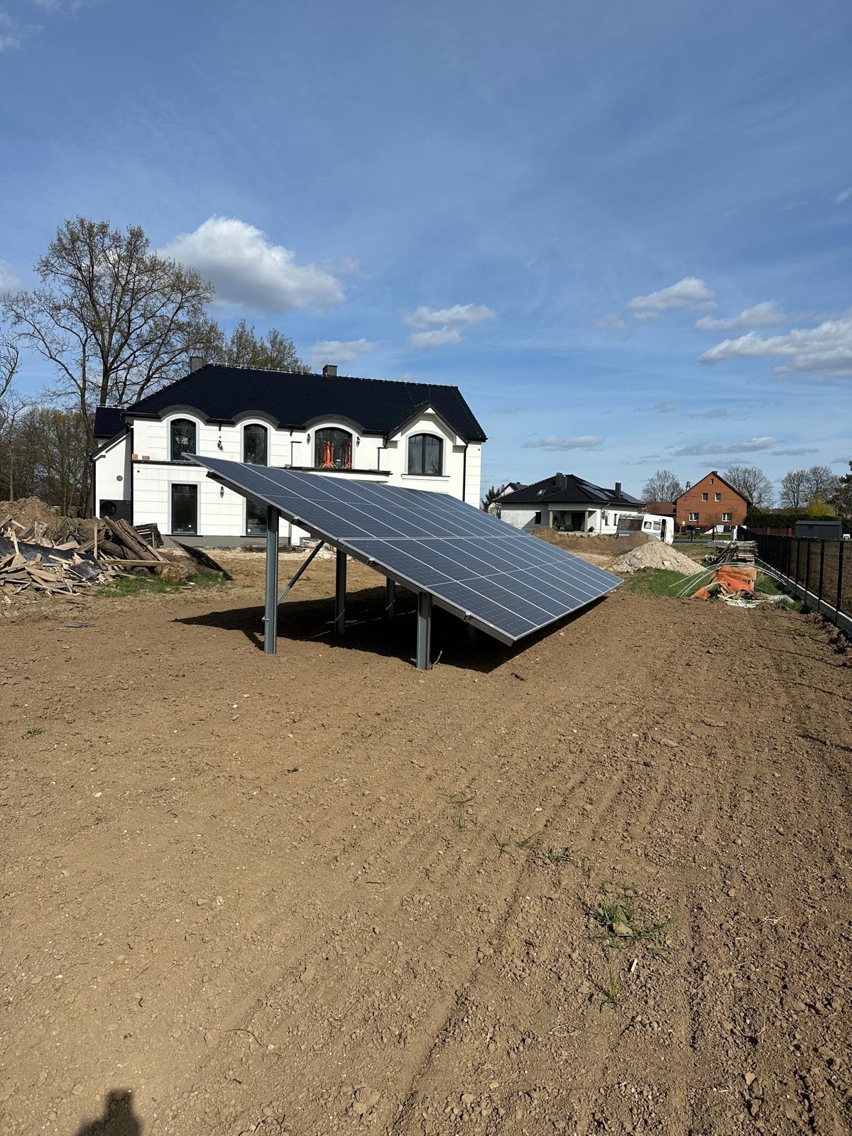Wolnostojąca instalacja fotowoltaiczna na przygotowanym gruncie, z widocznym domem jednorodzinnym w tle, w słoneczny dzień.