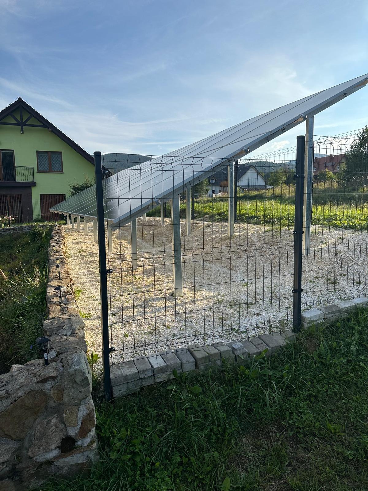 Instalacja paneli słonecznych na posesji, widok zza ogrodzenia z siatki. Panele zamontowane na metalowych słupach na gruncie wysypanym żwirem. W tle dom jednorodzinny i zabudowa wiejska.