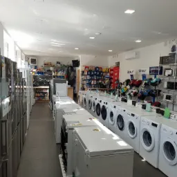 Wnętrze sklepu AGD z rzędem pralek automatycznych, zamrażarek skrzyniowych i lodówek, widoczne też półki z drobnym sprzętem AGD.