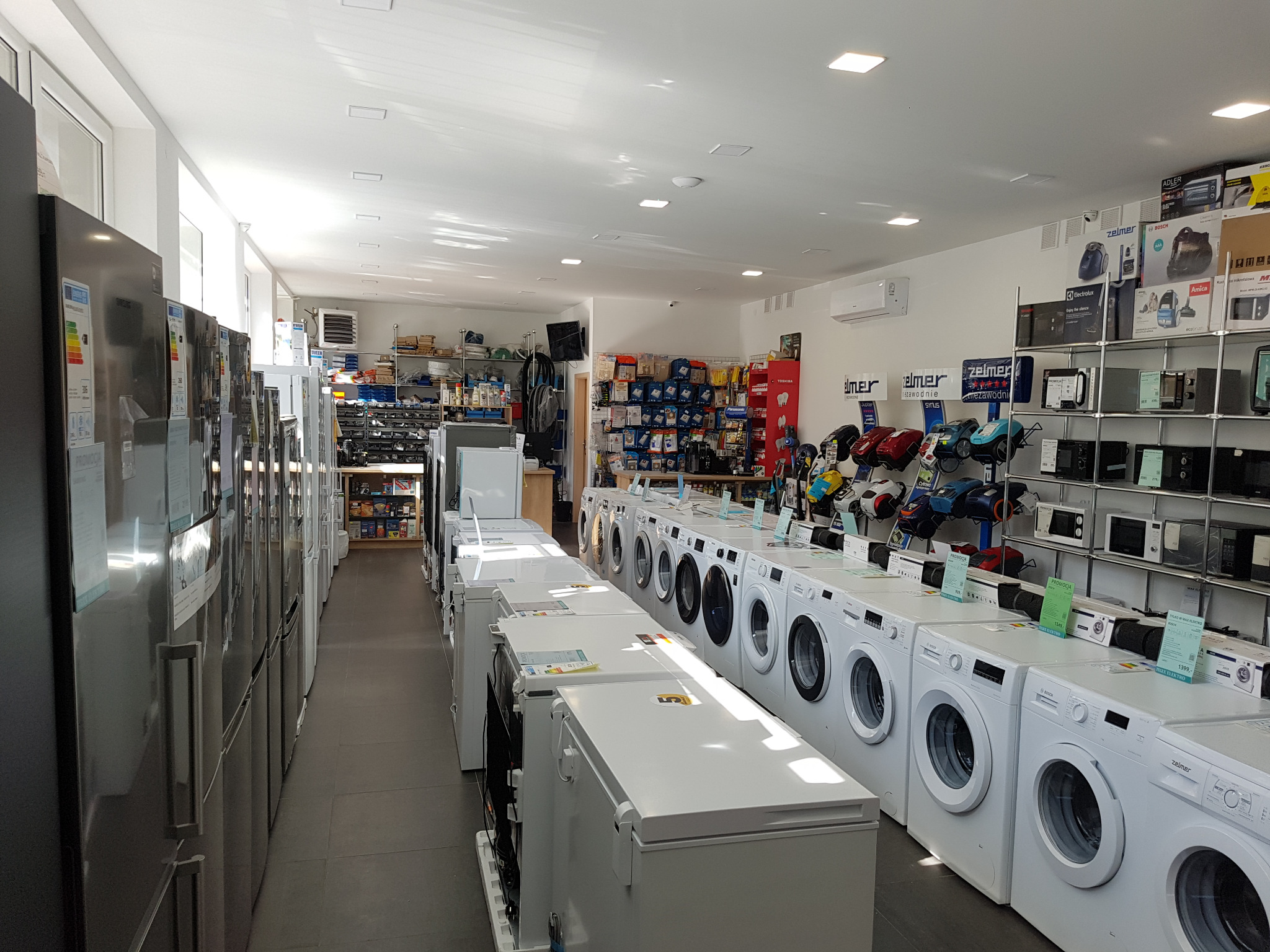 Wnętrze sklepu AGD z rzędem pralek automatycznych, zamrażarek skrzyniowych i lodówek, widoczne też półki z drobnym sprzętem AGD.