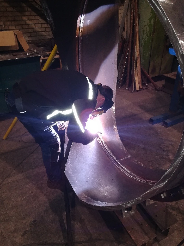 Pracownik w odzieży roboczej spawa element konstrukcji z metalu w warsztacie, widoczne iskry i spaw na okrągłym elemencie.