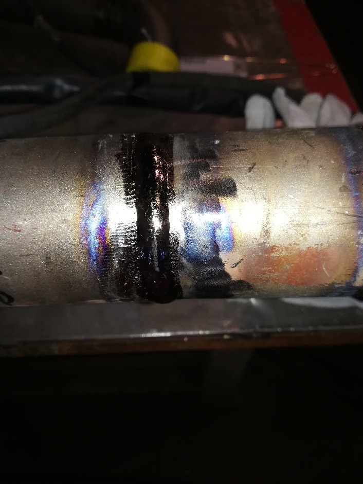 Spoina spawalnicza na metalowej rurze, widoczne przebarwienia termiczne i ślady oleju.