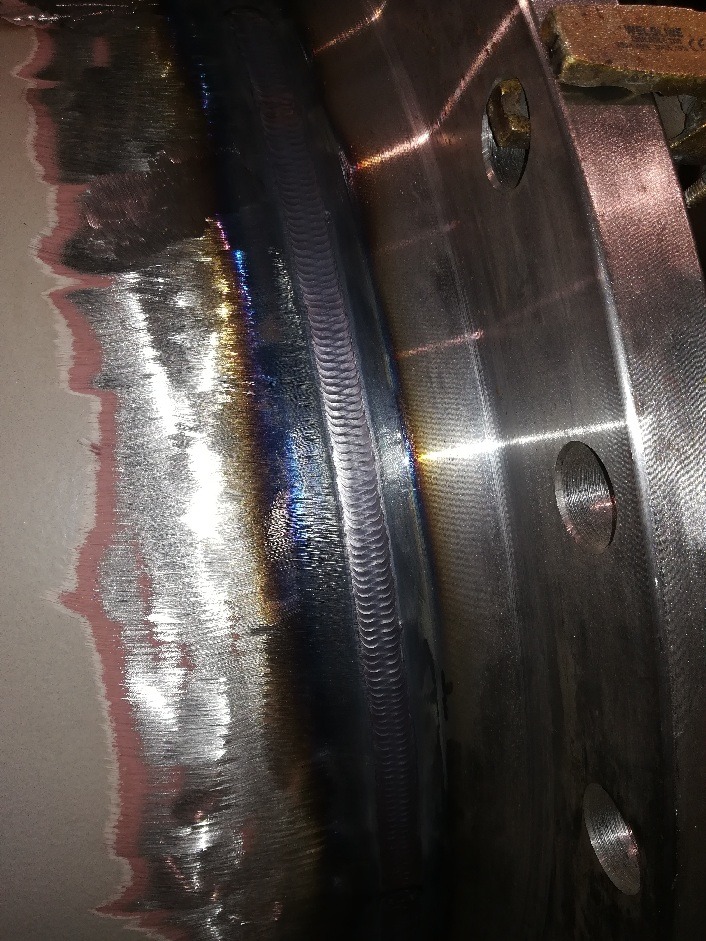 Błyszczące połączenie spawane dwóch metalowych elementów, widoczne ślady obróbki powierzchni i otwory montażowe.