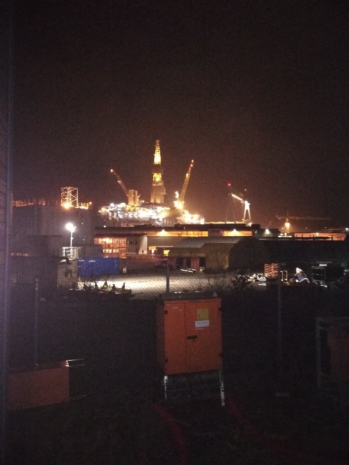 Nocny widok na platformę wiertniczą z dźwigami i oświetleniem, widzianą zza ogrodzenia i pomarańczowej szafki elektrycznej.