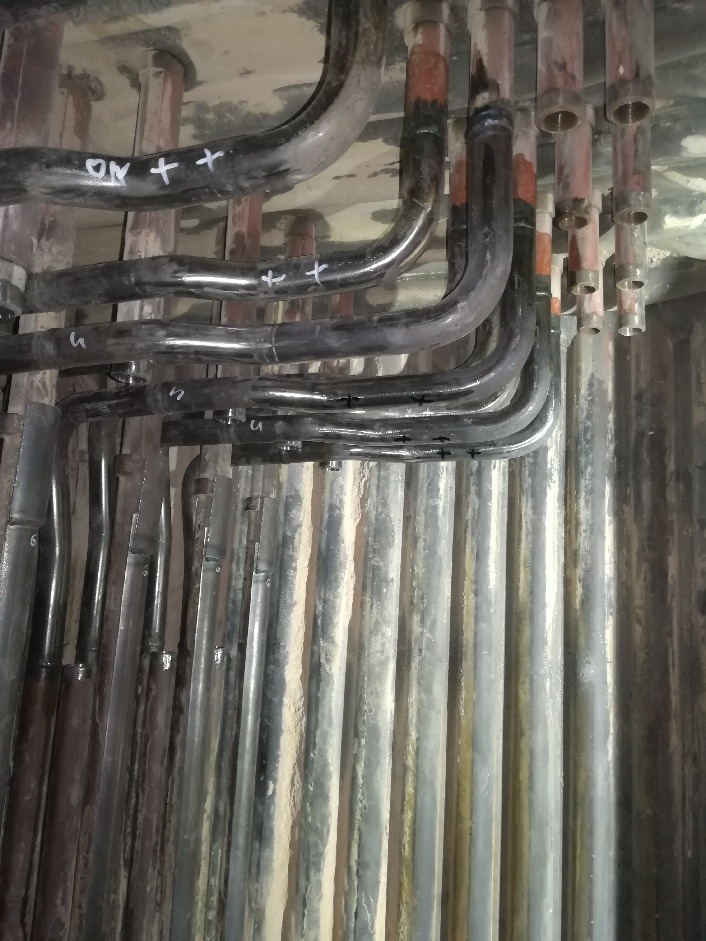 Zestaw rur metalowych z oznaczeniami, prawdopodobnie w kotłowni lub innym pomieszczeniu technicznym, widoczne ślady użytkowania i zabrudzenia.