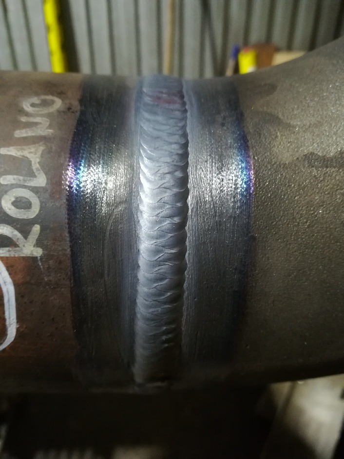 Szczegółowe zbliżenie na spaw na rurze, widoczne tęczowe przebarwienia termiczne i biały napis 'ROLLNO' na powierzchni.