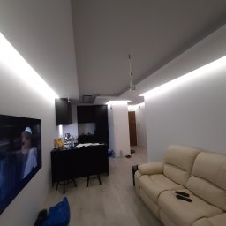 Dam-bud Damian Urbański - Nowoczesne wnętrze mieszkania po remoncie: białe ściany z oświetleniem LED, jasna podłoga, beżowa sofa, czarna zabudowa kuchenna z barkiem i telewizor na ścianie.