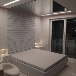 Dam-bud Damian Urbański - Nowoczesna sypialnia z białym łóżkiem, panelem ściennym za wezgłowiem i podwieszanym sufitem z oświetleniem LED, widok na okno balkonowe.