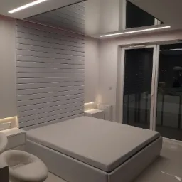 Nowoczesna sypialnia z białym łóżkiem, panelem ściennym za wezgłowiem i podwieszanym sufitem z oświetleniem LED, widok na okno balkonowe.