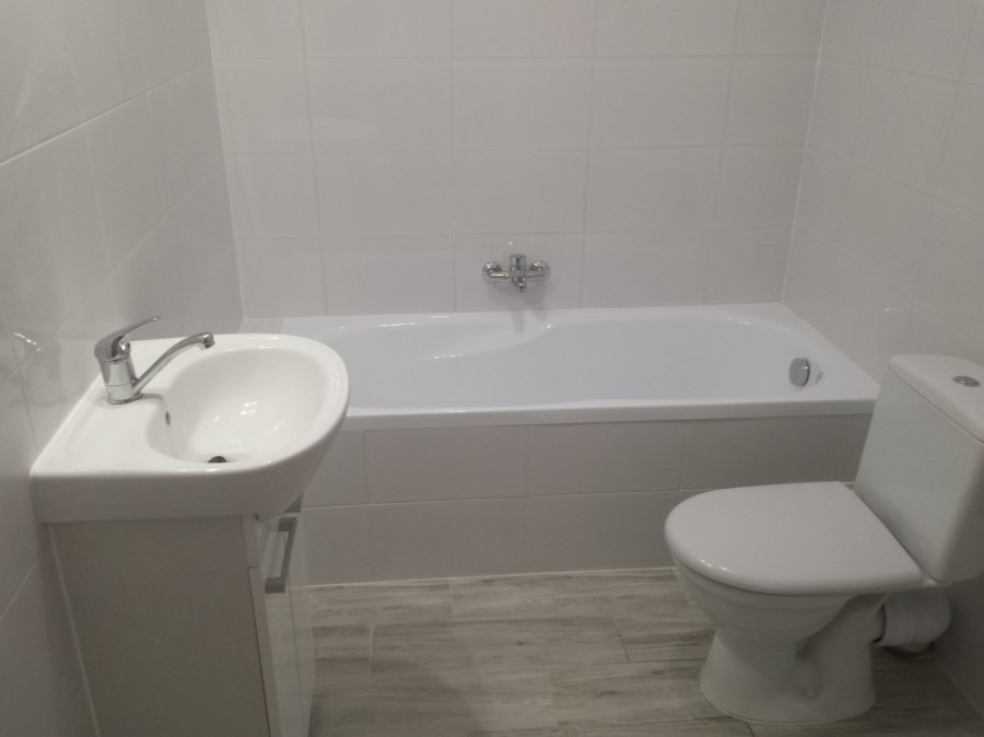 Nowoczesna, minimalistyczna łazienka z białą armaturą: umywalka z szafką, wanna obudowana płytkami i toaleta, wykończona szarymi płytkami imitującymi drewno.