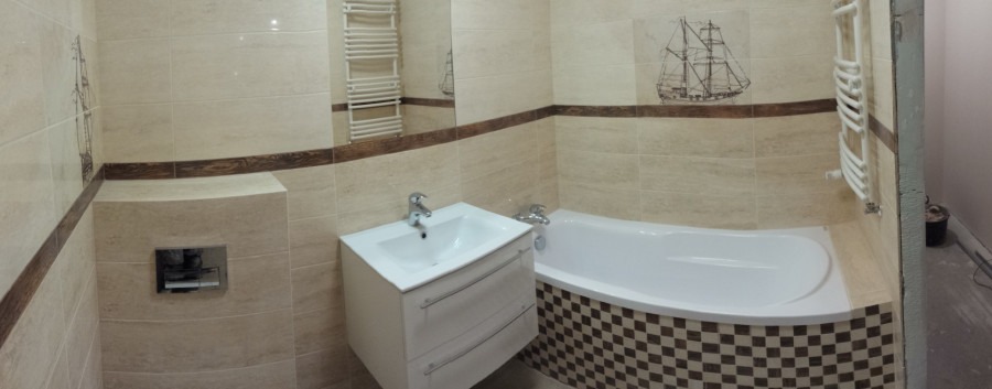 Wykończona łazienka z beżowymi płytkami, dekoracyjnym motywem żaglowca, białą umywalką z szafką i wanną obudowaną mozaiką w szachownicę, widoczny grzejnik drabinkowy i fragment niezakończonej ściany.