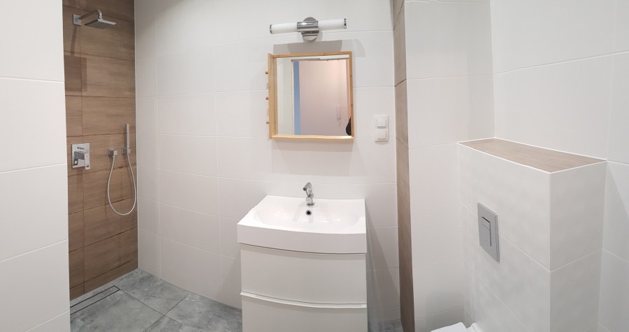 Nowoczesna łazienka z białymi płytkami, drewnianymi akcentami w strefie prysznica i lustrze, oraz białą umywalką z szafką i minimalistyczną armaturą.